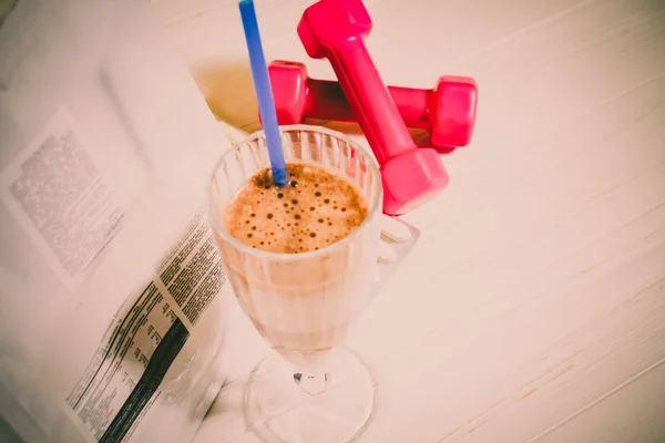 Egy proteinital pohárban, mellette rózsaszín súlyzók és táplálékkiegészítő csomag.